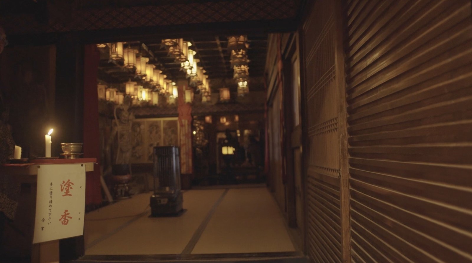An open temple hall, with a candle and warm light beneath hanging lamps.